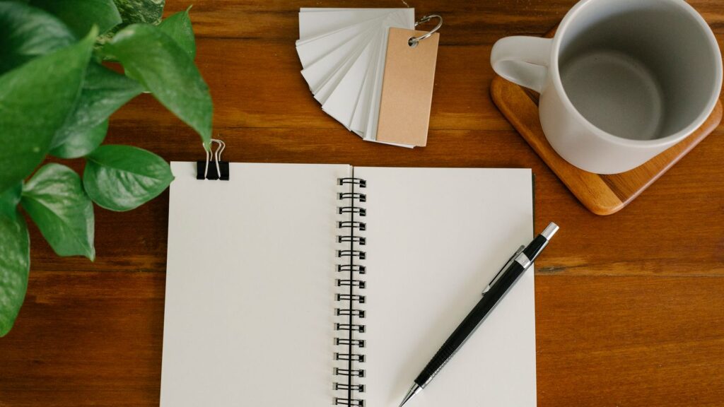 From above empty notebook with pen and tags placed near potted plant and empty mug on office table