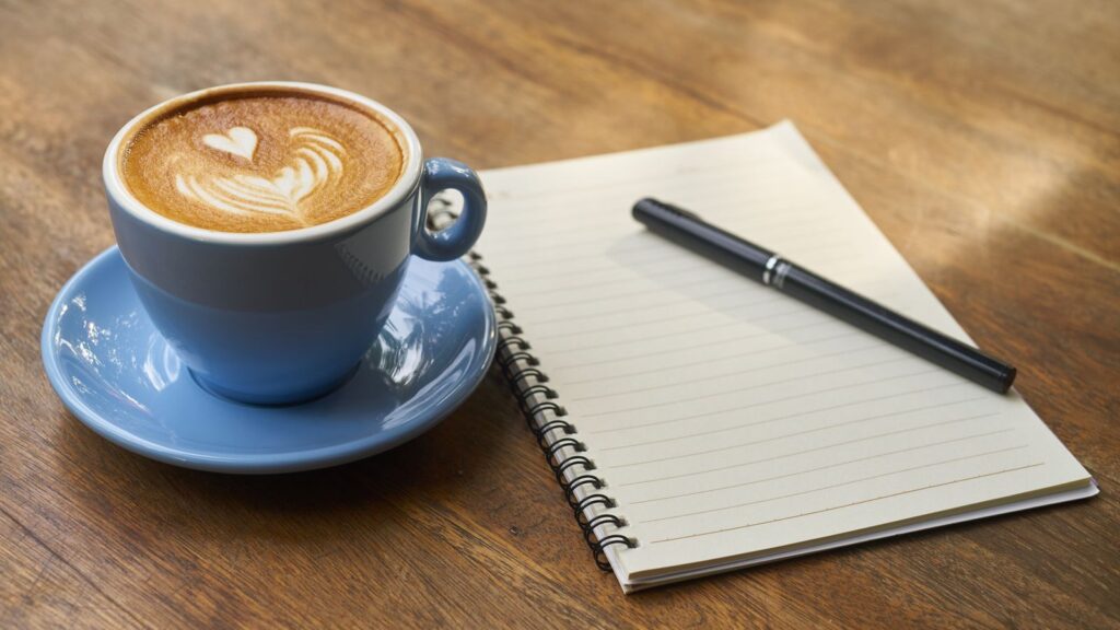 A calming scene of a latte with art in a blue cup beside a notebook and pen on a wooden table.