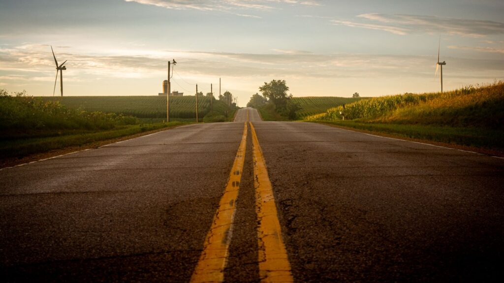 A scenic rural road flanked by wind turbines and fields under a summer sky.