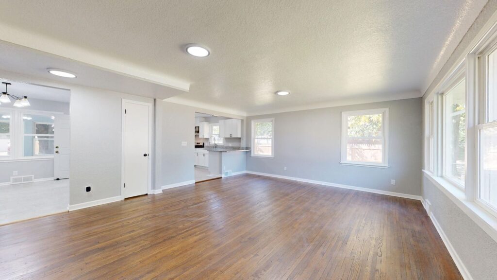 Sunlit empty living room with wooden flooring and large windows.